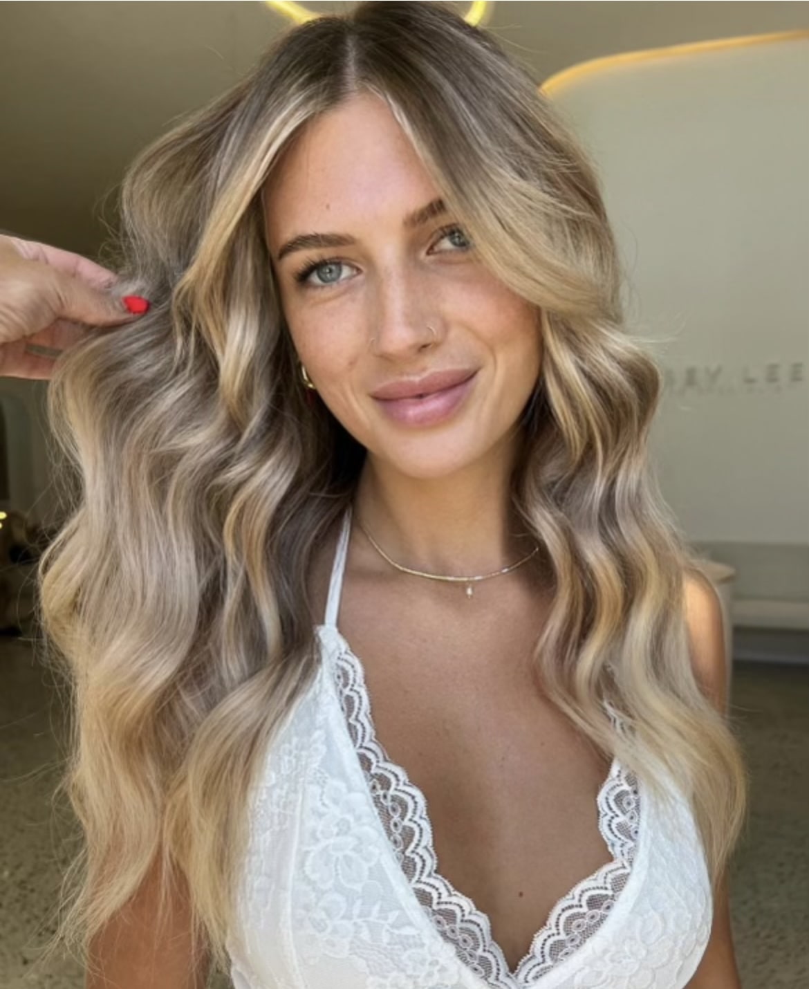 Smiling woman with wavy light blonde hair in a white top, showcasing the 'Dimensional Balayage' service at Kasey Lee Blonde Specialist.